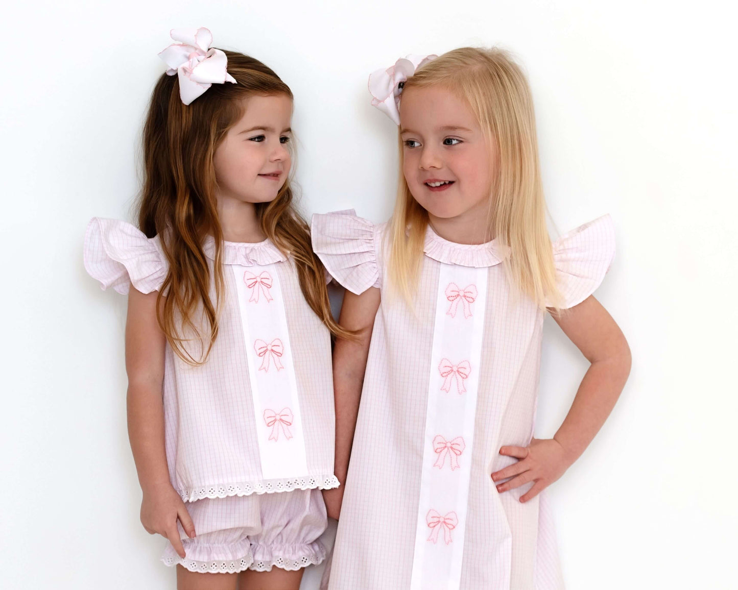 Two young girls wearing matching white dresses with pink bows against a white background