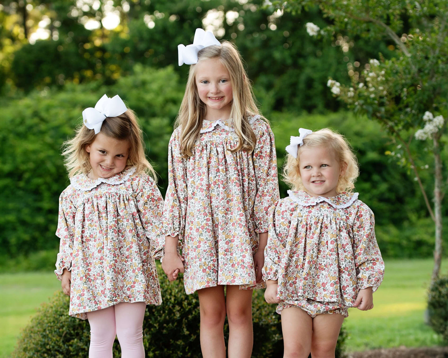 Three young girls in matching floral dresses standing outdoors with greenery in the background.