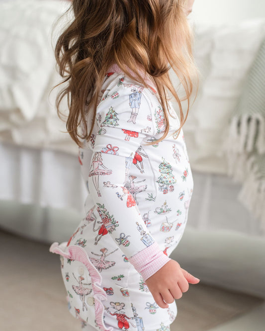 A child wearing a white long-sleeve pajama top with a colorful ballet dancer and holiday print, shown from the side in a softly lit bedroom setting.