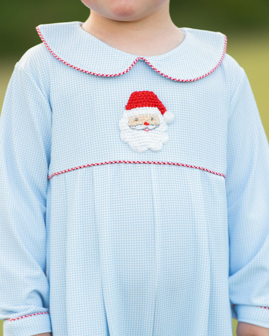 A close-up of a child wearing a light blue gingham dress with a red and white braided Peter Pan collar, a red and white braided horizontal trim line, and a central Santa Claus face applique.