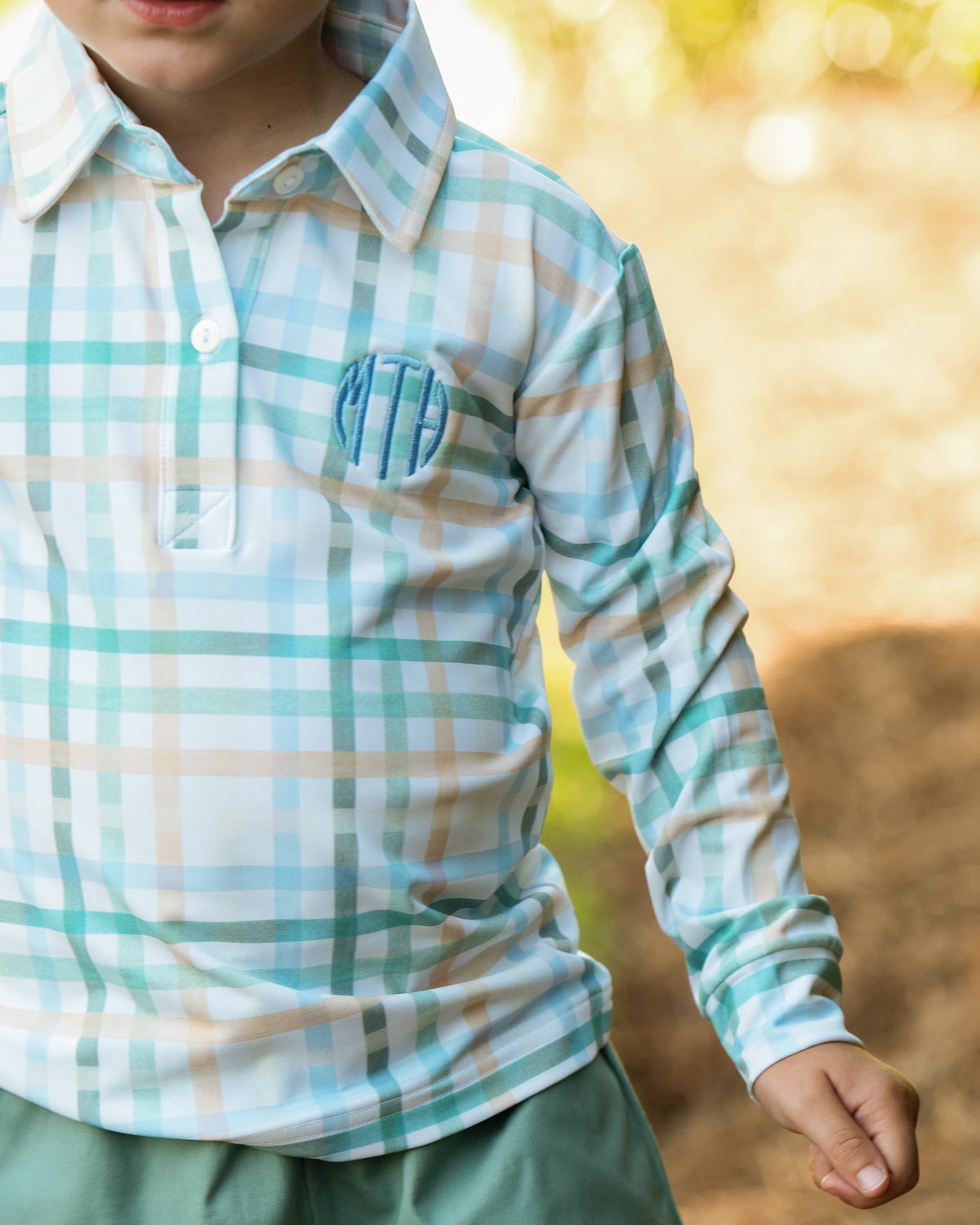 A close-up of a child wearing a long-sleeve plaid polo shirt in white, light blue, and beige, with a blue monogram on the chest, set against a blurred outdoor background.