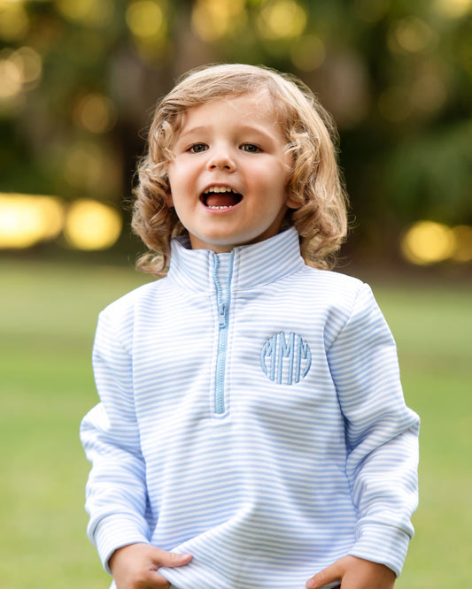 A little boy with blond hair standing outside with a blurry background, wearing a blue striped knit pullover with a quarter-zipper and a blue round monogram