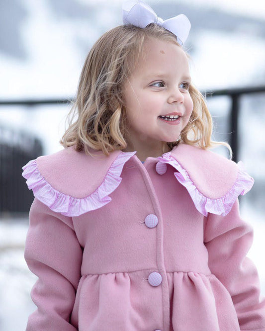 A little girl standing outside with a white bow in her hair, wearing a pink wool coat trimmed in pink gingham and gingham buttons