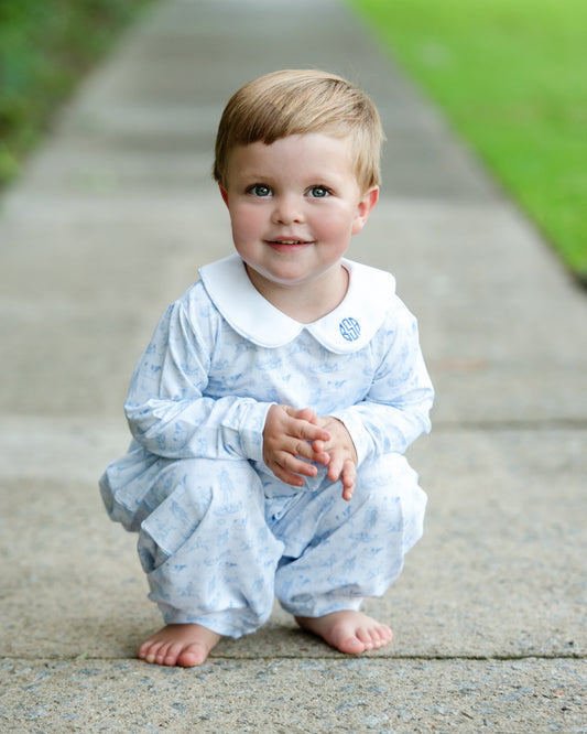 A toddler crouching barefoot on a concrete path outdoors, wearing a light blue patterned long-sleeve pajama set with a white Peter Pan collar and a small blue crest on the collar.