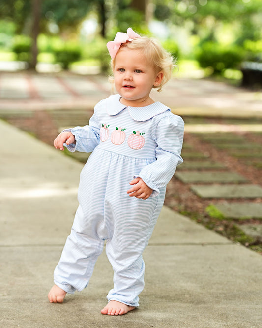 A toddler girl walking barefoot on an outdoor concrete path, wearing a light blue and white striped long-sleeve romper with embroidered peach details on the chest and a pink bow in her blonde hair.