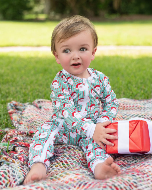 A toddler sitting on a colorful blanket on a grassy lawn, wearing a green and white Santa Claus patterned pajama set and holding a red wrapped gift box.