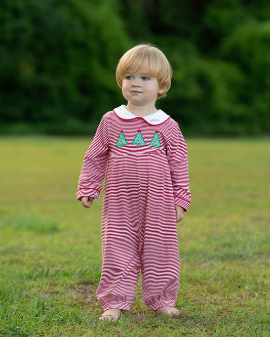 A toddler standing outdoors on grass wearing a pink and white striped footless one-piece jumpsuit with a white Peter Pan collar and three green embroidered trees on the chest.