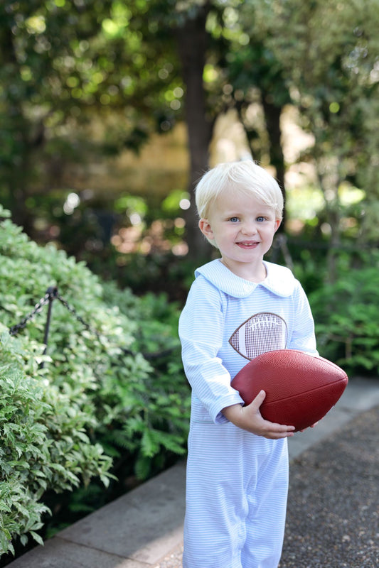 A toddler with blonde hair stands outdoors in a garden setting, wearing a light blue and white striped footed onesie with a football graphic on the chest and holding a red football.
