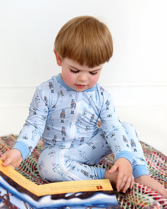 A toddler with light brown hair sits on a colorful woven blanket, reading a picture book. The child is wearing a light blue long-sleeve zip-up pajama set with a small repeating astronaut pattern.