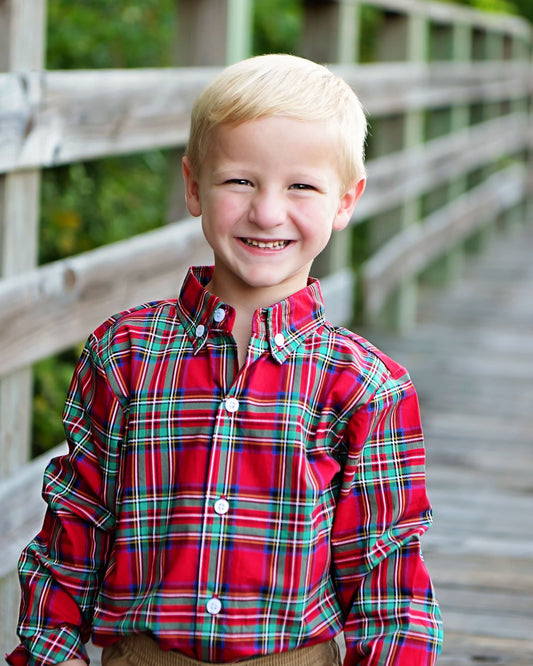 A young blond boy smiling while wearing a red plaid button-down shirt with green and blue lines, standing on a wooden boardwalk.