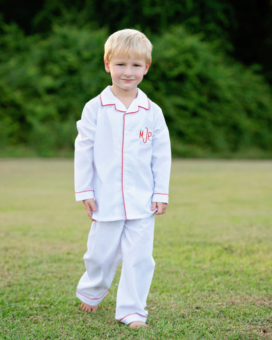 A young blond boy wearing a white long-sleeve pajama set with red piping and a red monogram on the chest, standing barefoot on a grassy lawn with a blurred green background.