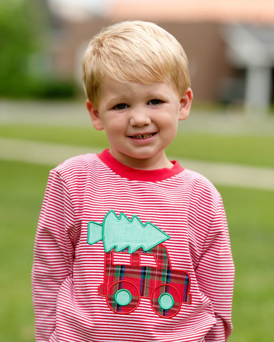 A young blonde boy smiling outdoors, wearing a red and white striped long-sleeve shirt with a red truck applique carrying a light green Christmas tree.