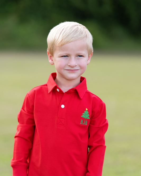 A young blonde boy wearing a bright red long-sleeve polo shirt with a green embroidered Christmas tree and monogram logo on the left chest, standing outdoors on a blurred green lawn.