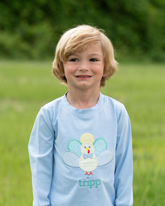 A young blonde boy wearing a light blue long-sleeve shirt with a turkey applique design and the name 'tripp' printed below it, standing in a grassy outdoor field.