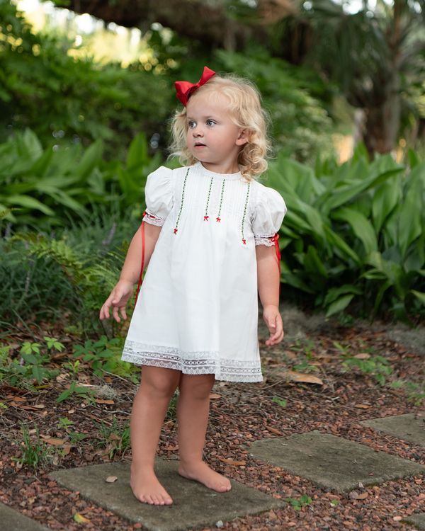 A young blonde girl wearing a white short-sleeve dress with red ribbon accents and a red bow in her hair, standing barefoot on a stone path in a lush green garden.
