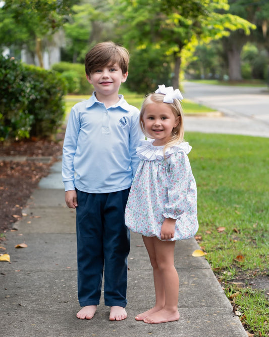 A young boy and girl standing barefoot on a sidewalk outdoors. The boy wears a light blue long-sleeve polo shirt with dark navy pants, and the girl wears a white floral dress with a ruffled collar and a white bow in her hair.
