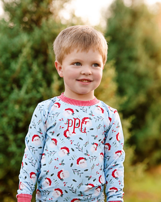 A young boy smiling outdoors while wearing a light blue long-sleeve pajama shirt with a Santa Claus print and red gingham trim. The shirt features the monogram 'PPG' in red on the chest.