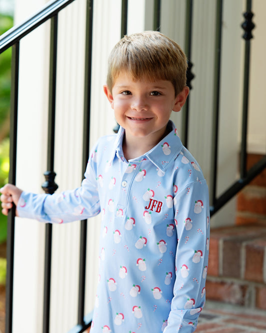 A young boy smiling while wearing a light blue long-sleeve collared pajama shirt with a white Santa Claus pattern and a personalized red monogram 'JFB' on the chest, standing in front of an outdoor staircase.