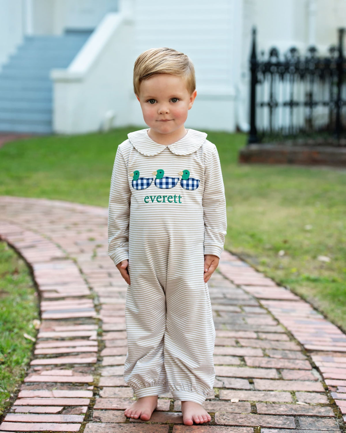 A young boy stands barefoot on a brick path outdoors, wearing a cream and white striped long-sleeve romper with a white Peter Pan collar. The front features a blue and white gingham crocodile graphic with the name 'everett' and three green alligator heads embroidered below it.