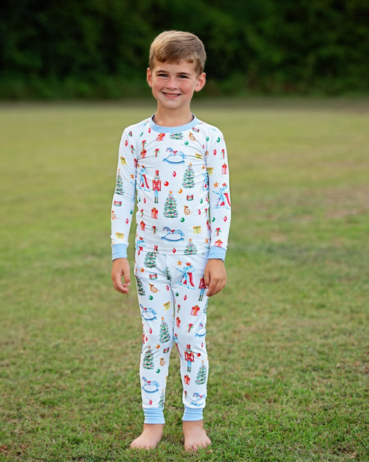 A young boy stands barefoot on a grassy lawn wearing a light blue long-sleeve pajama set with a colorful Christmas-themed print of trees, gifts, and snowman motifs.