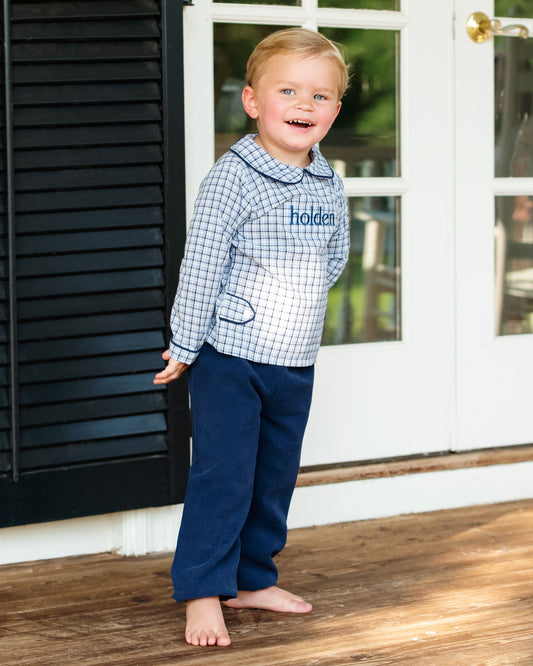 A young boy stands barefoot on a wooden porch wearing a light blue and white gingham long-sleeve pajama top with 'helden' embroidered on the chest, paired with navy blue pants.