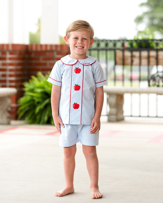 A young boy stands barefoot outdoors wearing a light blue short-sleeve collared shirt and matching shorts. The shirt has red trim and three red apple graphics on the front, with the name 'TERRIE' printed above the first apple.