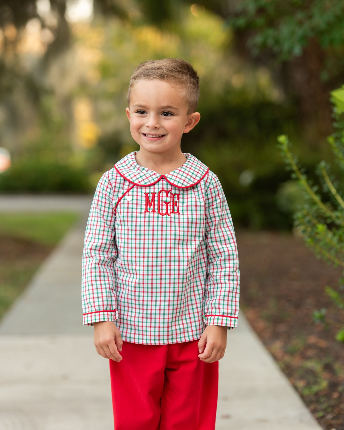 A young boy stands on a paved path outdoors wearing a long-sleeve red, green, and white gingham shirt with red trim and the letters 'MGE' embroidered in red on the chest, paired with bright red pants.