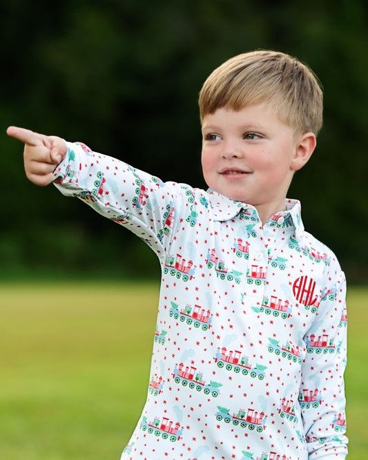 A young boy wearing a white long-sleeve button-up shirt with a red and green train pattern and a red monogram on the chest, pointing outdoors toward the left against a blurred green background.