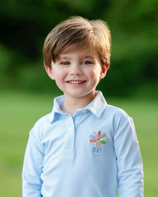 A young boy with light brown hair smiling outdoors, wearing a light blue long-sleeve polo shirt with a colorful embroidered logo on the chest.