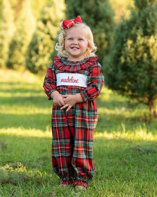 A young child wearing a red and green plaid jumpsuit with a white name patch that reads 'madeline' in red script, standing outdoors on grass with trees in the background.