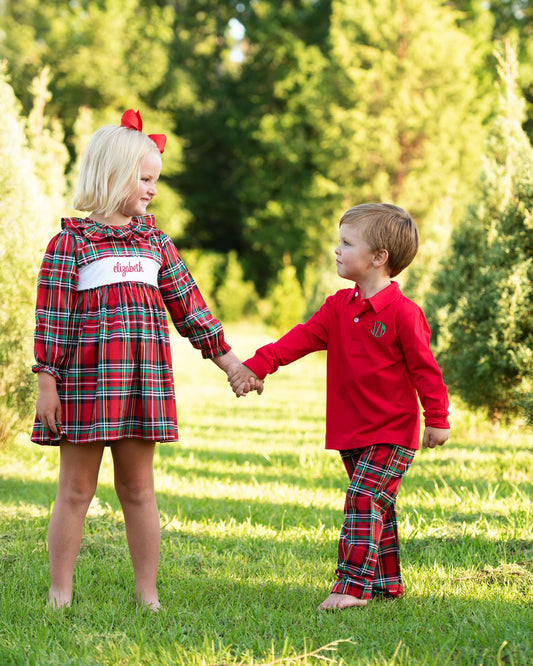 A young girl and boy holding hands outdoors on a grassy lawn. The girl wears a red plaid long-sleeve dress with a white name tape reading 'dixie' and a red bow in her hair.