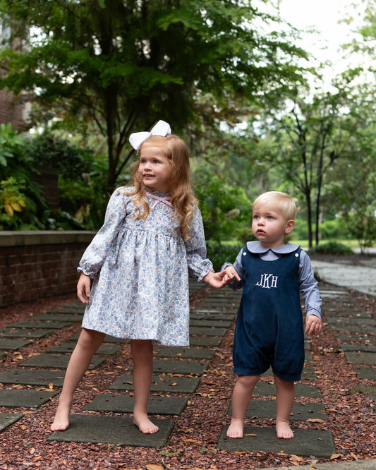 A young girl in a floral long-sleeve dress with a white bow and a toddler in navy blue overalls, holding hands on a stone path in a garden.