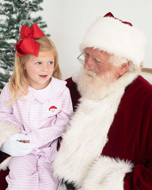 A young girl in pink and white striped pajamas with a large red bow sits next to Santa Claus. The girl has white gloves and a small Santa-themed patch on her pajama chest.