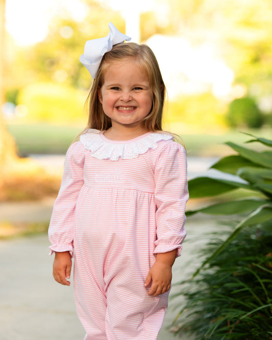 A young girl smiling outdoors while wearing a pink and white striped long-sleeve romper with a white ruffled collar and a white bow in her hair.