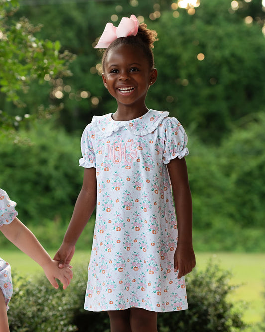 A young girl smiling outdoors while wearing a white short-sleeve dress with a colorful floral print and a ruffled Peter Pan collar. She has a pink bow in her hair, and another person is partially visible holding her hand on the left.