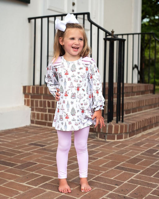 A young girl stands barefoot on a brick porch wearing a long-sleeve white tunic dress with a colorful winter tree and snowman print, paired with light pink leggings and a white bow in her hair.