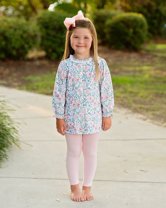 A young girl stands barefoot on a concrete path outdoors, wearing a long-sleeve floral tunic top with a high ruffled collar and light pink leggings. She has a pink bow in her hair.