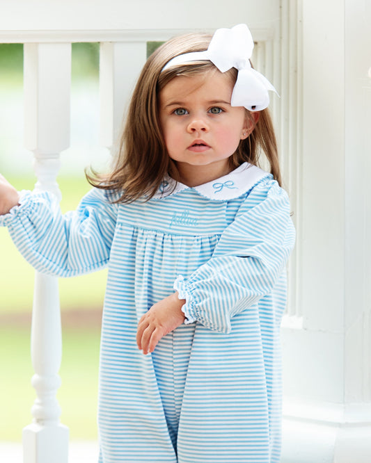 A young girl wearing a light blue and white striped long-sleeve nightgown with a white collar and a large white bow headband, standing on a porch.
