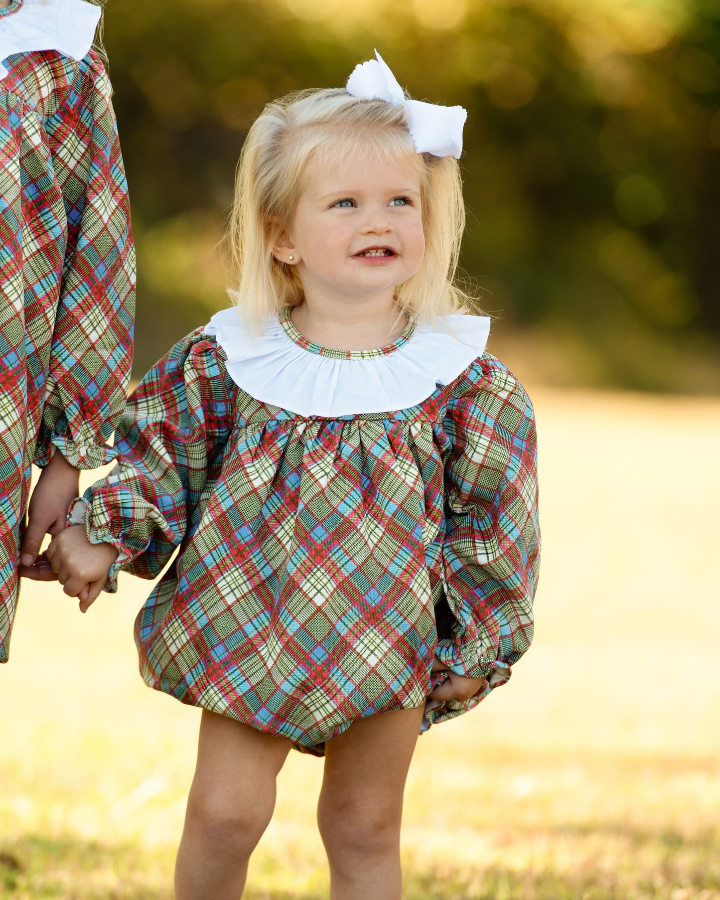 A young girl wearing a plaid bubble romper with a white ruffled collar and a white hair bow, standing outdoors in a grassy field.