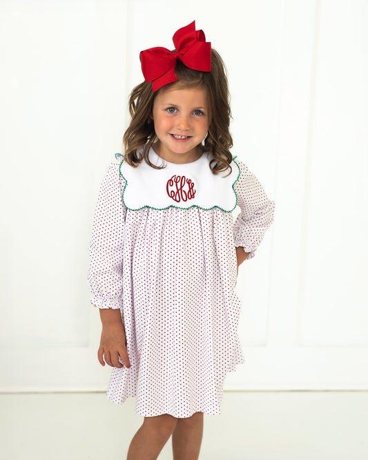 A young girl wearing a white long-sleeve dress with small red polka dots, a white bib with green trim and red monogram lettering, and a large red bow in her hair, standing against a white paneled background.