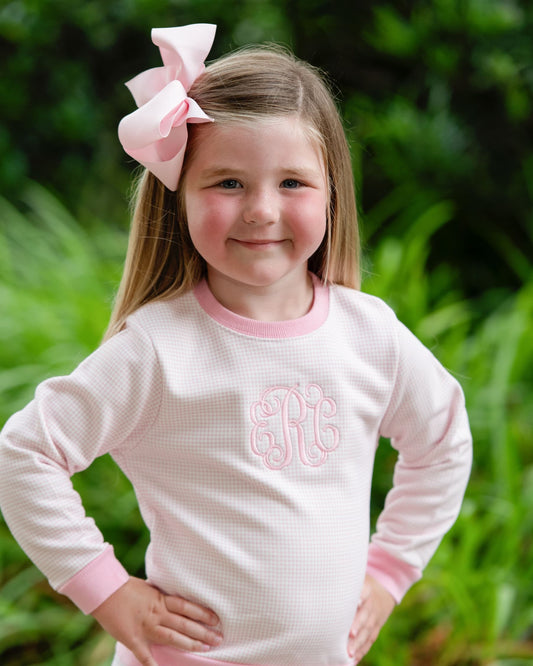 A young girl with a large pink bow in her hair wears a light pink waffle-knit long-sleeve shirt with a pink collar and cuffs, featuring a pink monogram on the chest, set against a blurred green background.
