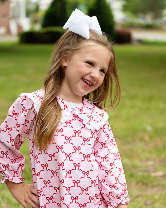 A young girl with a large white bow in her hair is smiling outdoors on a grassy lawn. She is wearing a light pink long-sleeve dress with a repeating red bow pattern and a white ruffled collar.
