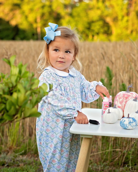 A young girl with blonde hair and a blue bow stands outdoors next to a small white table, wearing a light blue floral long-sleeve dress. Decorative pumpkins are on the table, with a blurred field and trees in the background.