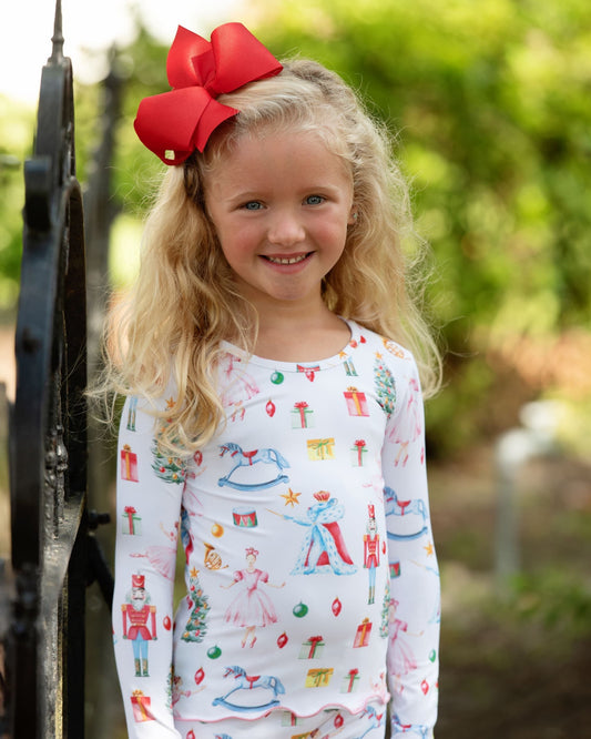 A young girl with blonde hair and a large red bow smiles while wearing a white long-sleeve pajama top with a colorful nutcracker and toy print, standing outdoors next to a black gate.