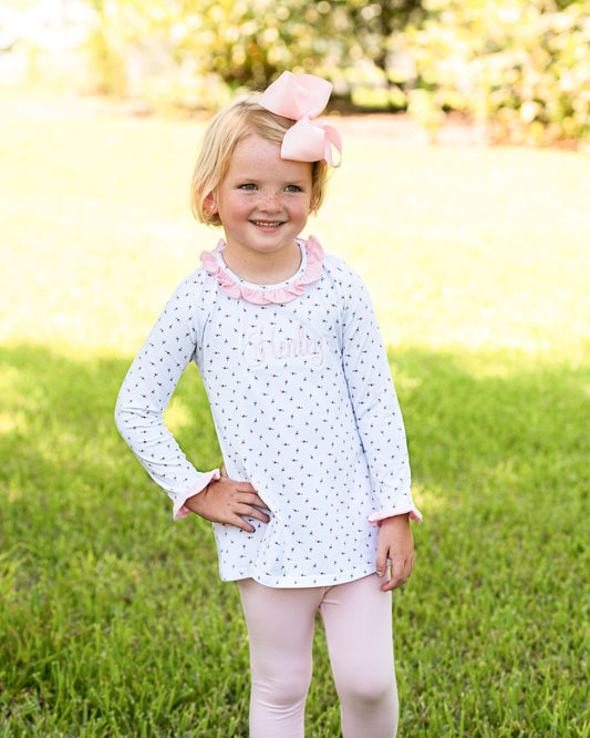A young girl with blonde hair and a pink bow stands on a green lawn wearing a white long-sleeve tunic top with a pink ruffled collar and small pink polka dots, paired with light pink leggings.