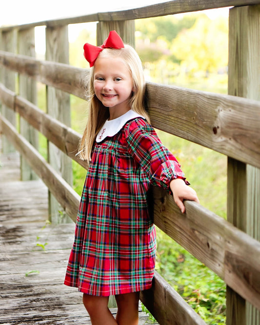 A young girl with blonde hair and a red bow stands on a wooden boardwalk wearing a red, green, and blue plaid dress with a white collar.