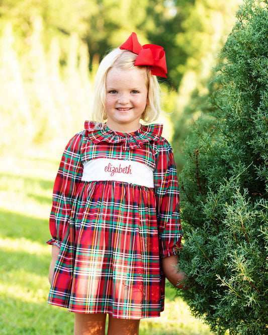 A young girl with blonde hair and a red bow stands outdoors next to a green tree, wearing a red plaid dress with a white chest panel embroidered with the name 'elizabeth' in red script.