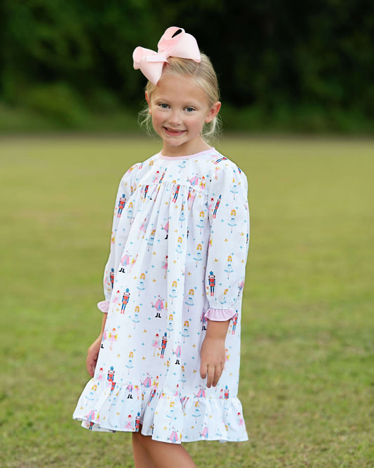 A young girl with blonde hair in a large pink bow stands outdoors on a grassy lawn wearing a white long-sleeve dress with a colorful nutcracker and fairy print.