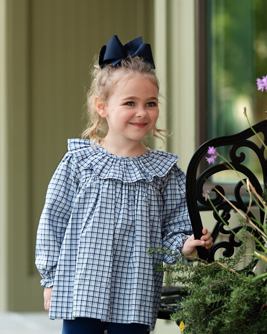 A young girl with blonde hair in a navy bow stands on a porch wearing a blue and white gingham long-sleeve top with a ruffled collar and dark blue pants.