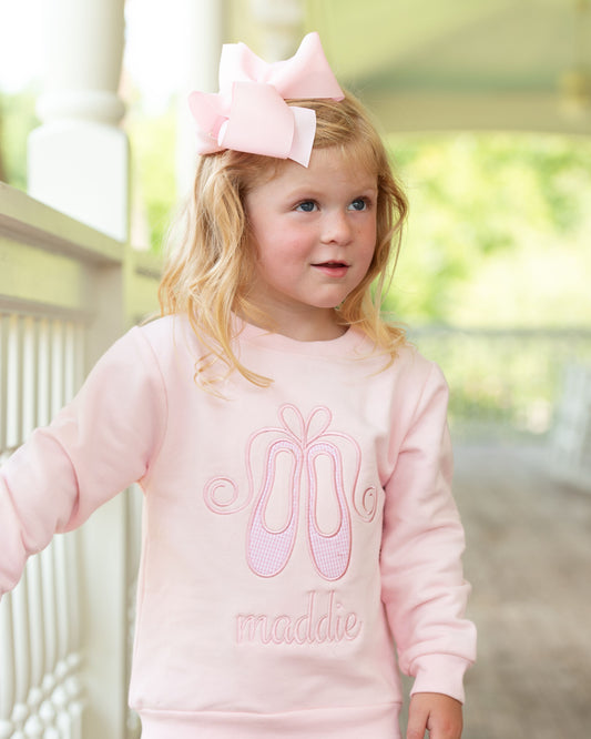 A young girl with blonde hair wearing a large light pink bow in her hair and a matching light pink sweatshirt with a ballet shoe graphic and the name 'maddie' embroidered on the front, standing on a porch.